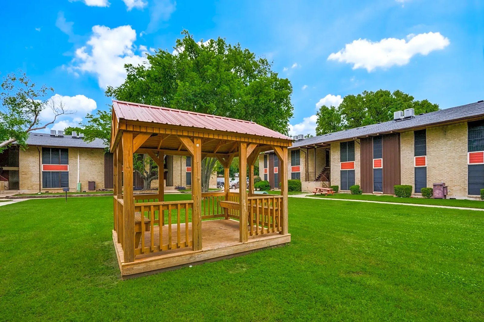 Grassy courtyard with a wooden gazebo, mature trees and two-story brick buildings